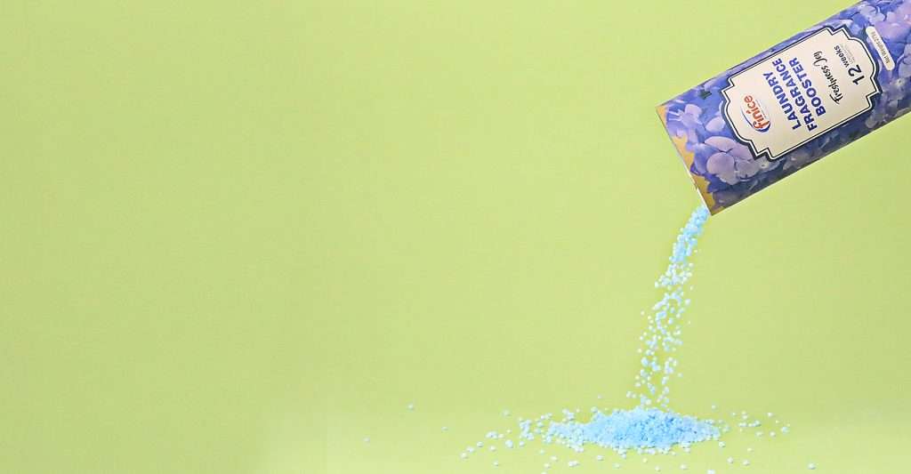 A container of blue laundry fragrance booster beads pouring onto a surface against a green background, with the product labeled "Laundry Fragrance Booster" and featuring floral design packaging.